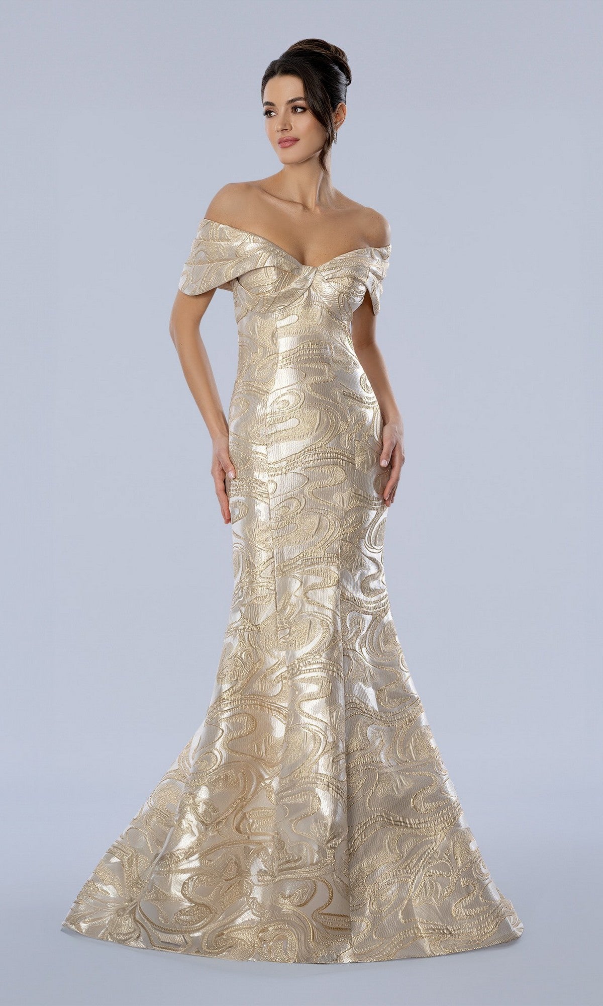 A woman stands against a plain light background wearing the Stella Couture 26360 long formal dress in gold, an off-the-shoulder evening gown crafted from shimmering metallic brocade, featuring a fitted bodice and a flared mermaid skirt.